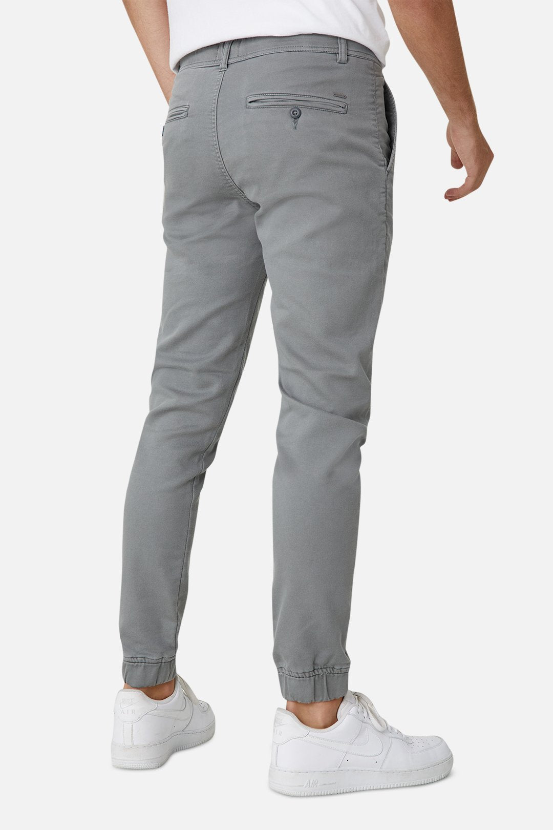 A person is shown from the back wearing Industrie The Drifter Chino Pant in gray, featuring a slim fit jogger style with an elastic waist and welt pockets, paired with simple white sneakers. The background is plain white.