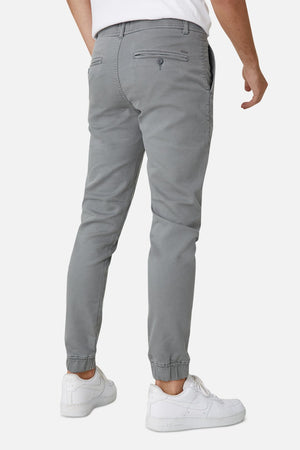 A person is shown from the back wearing Industrie The Drifter Chino Pant in gray, featuring a slim fit jogger style with an elastic waist and welt pockets, paired with simple white sneakers. The background is plain white.