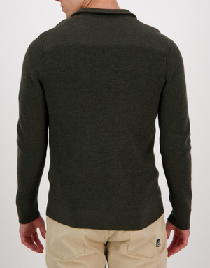 A person is displaying the back of their Swanndri Magnet Bay v2 Merino Jumper, paired with beige pants. The dark green jumper, crafted from premium Merino wool, subtly contrasts with the plain light background.