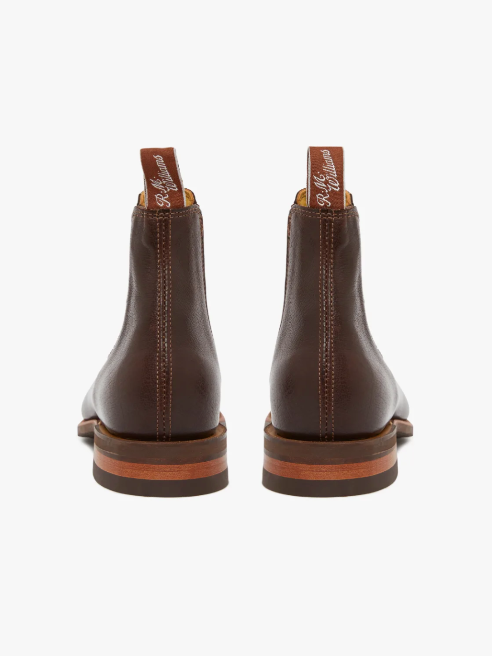The RM Williams Comfort Kangaroo Craftsman Boot, viewed from the back, showcases its brown leather finish and is handcrafted in Australia. It features pull tabs with white text and stacked heels complemented by wood detailing. Vertical stitching runs along the heel, and they come equipped with a comfort innersole. The background is white.