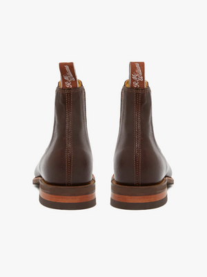 The RM Williams Comfort Kangaroo Craftsman Boot, viewed from the back, showcases its brown leather finish and is handcrafted in Australia. It features pull tabs with white text and stacked heels complemented by wood detailing. Vertical stitching runs along the heel, and they come equipped with a comfort innersole. The background is white.
