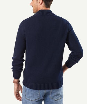 A person facing away wears a navy blue Gazman Chunky Button Neck Knit with a high collar and light blue jeans. The cotton sweater in regular fit has ribbed cuffs and hem against a plain, light background.