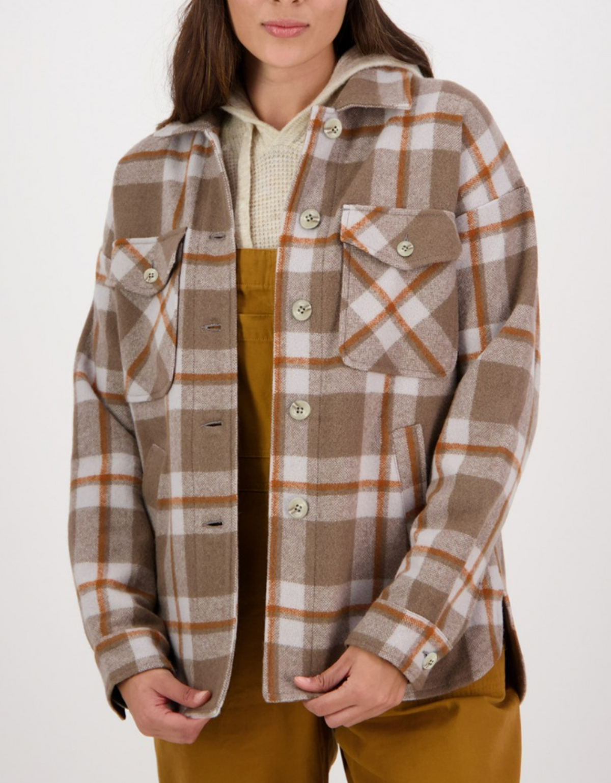 A person wearing a Swanndri Anchorage Wool Shirt Jacket in brown and white plaid over a beige knitted Merino wool sweater and brown overalls. The shacket features two front pockets with buttons and buttoned cuffs. The person's hands are loosely clasped in front, set against a plain white background.