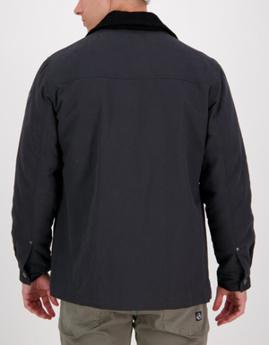 A person wears the Swanndri Barrytown v2 Canvas Work Jacket, which is dark with a black collar and buttoned cuffs, over light pants. The jacket has a quilted lining for added warmth and features a subtle logo on the lower back. They face away from the camera against a plain white background.