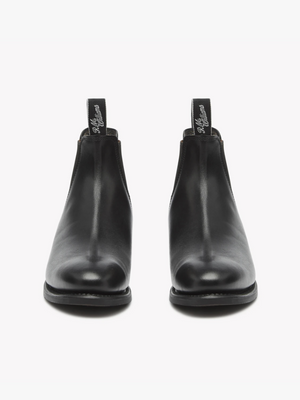 The RM Williams Adelaide Rubber Sole Boot is a pair of black leather Chelsea boots with a smooth finish. Crafted from Yearling leather, these classic boots feature elastic side panels and fabric pull tabs at the back with embroidered text. They are positioned facing forward on a plain white background.
