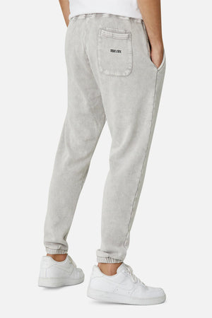 The person is donning Industrie The Del Sur Track Pant in light gray, featuring a back pocket with an "ITBO'S SPACE" label and an elastic drawstring waist. Paired with white sneakers, one hand rests casually in a front pocket against a plain white background.