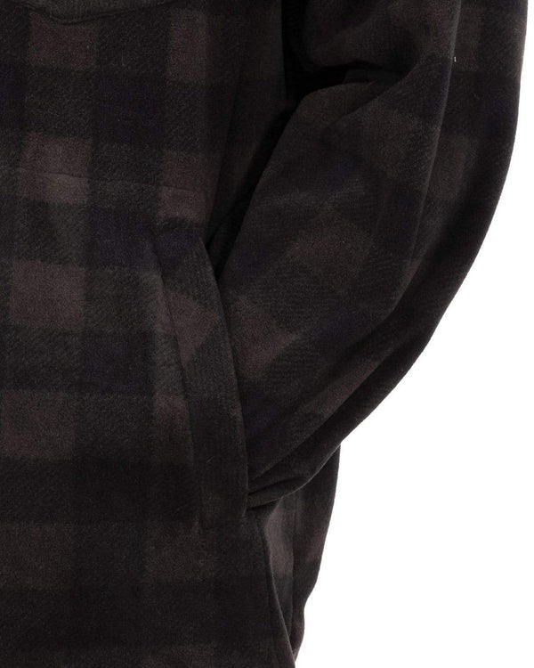 A close-up shows the Outback Mens Big Shirt, a dark plaid mid-weight with 100% polyester fabric in a black and brown checkered pattern. Only the garment and a partial arm are visible, with one hand tucked into a side pocket.