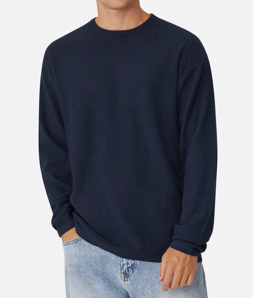 A person wearing Industrie The Aries Knit, a dark navy blue, long-sleeve textured sweater with an oversized fit, paired with light blue jeans. The 100% cotton sweater features a crew neck collar, and the individual is posing casually with one hand in their pocket against a plain white background. The person's face is not visible.