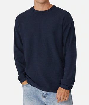 A person wearing Industrie The Aries Knit, a dark navy blue, long-sleeve textured sweater with an oversized fit, paired with light blue jeans. The 100% cotton sweater features a crew neck collar, and the individual is posing casually with one hand in their pocket against a plain white background. The person's face is not visible.