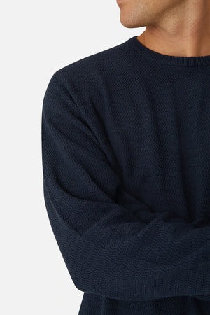 A person in an oversized navy blue textured sweater, named "Industrie The Aries Knit" and made from 100% cotton, stands with arms crossed. They look to the side against a plain light gray backdrop, highlighting their upper body and part of their face.