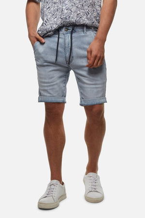 Wearing Industrie The Drifter Denim Short in light blue, a patterned shirt, and white sneakers, the person stands against a plain background. With one hand in their pocket, they exude an effortlessly casual vibe.