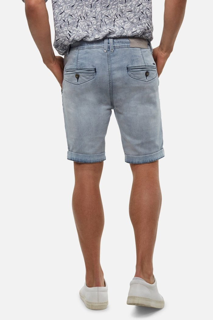 A person in Industrie The Drifter Denim Short, featuring light denim with rolled cuffs and two buttoned back flap pockets, paired with a patterned short-sleeve shirt and white sneakers, stands facing away against a neutral background.