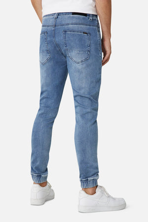 A person in stylish Industrie The Drifter Denim Pant, light blue with a slim fit and elastic ankle cuffs, stands back to the camera wearing white sneakers. Visible back pockets complete the comfortable look.