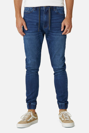Wearing Industrie The Drifter Denim Pant, a person stands against a plain white backdrop. The blue denim joggers with a relaxed fit and drawstring waistband are perfectly paired with a white shirt and beige sneakers featuring white laces and soles.
