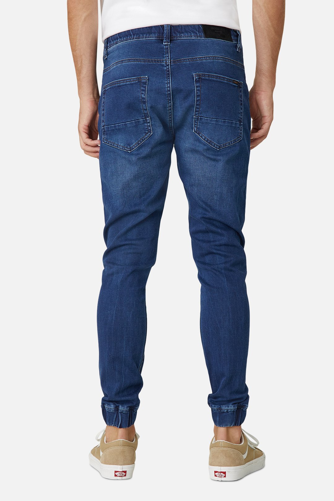 A person is shown from the back wearing Industrie The Drifter Denim Pant in dark blue with a relaxed fit and beige sneakers. The jeans feature two back pockets and folded cuffs, paired with a crisp white shirt, set against a plain white background for an effortlessly stylish look.