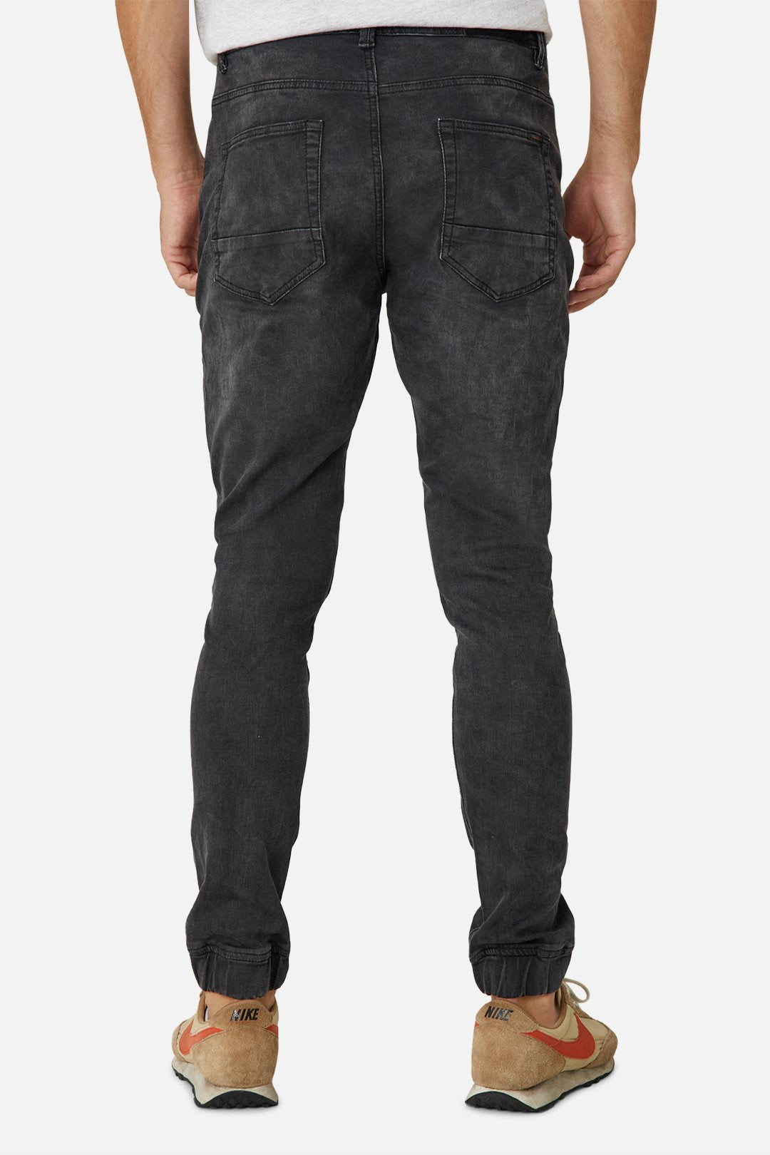 A person wearing stylish, Industrie The Drifter Denim Pant in faded black and beige Nike sneakers is shown from the back against a plain white background. The slightly fitted jeans are gathered at the ankles, and the sneakers feature red swooshes. The right shoe is partially untied.