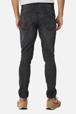 A person wearing stylish, Industrie The Drifter Denim Pant in faded black and beige Nike sneakers is shown from the back against a plain white background. The slightly fitted jeans are gathered at the ankles, and the sneakers feature red swooshes. The right shoe is partially untied.