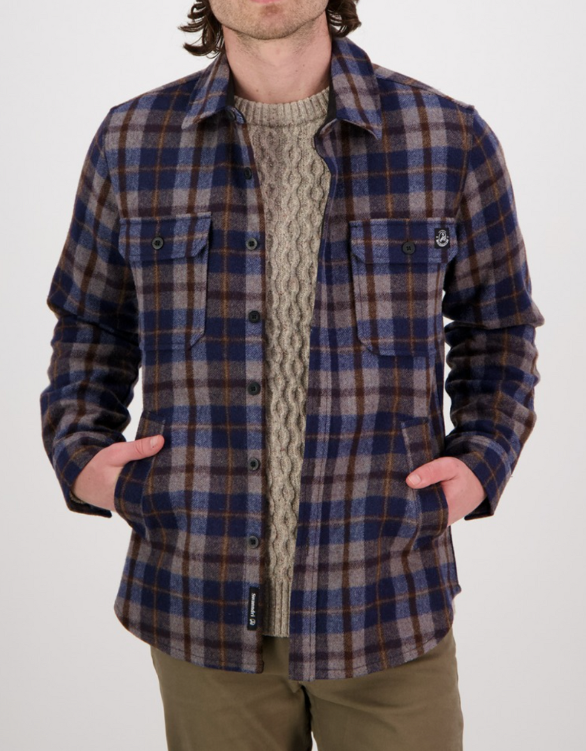 A person wearing a blue and brown plaid Swanndri Kiaraki v3 Wool Shacket over a beige cable-knit sweater, hands in pockets, stands against a plain white background.