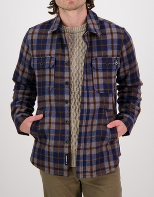 A person wearing a blue and brown plaid Swanndri Kiaraki v3 Wool Shacket over a beige cable-knit sweater, hands in pockets, stands against a plain white background.