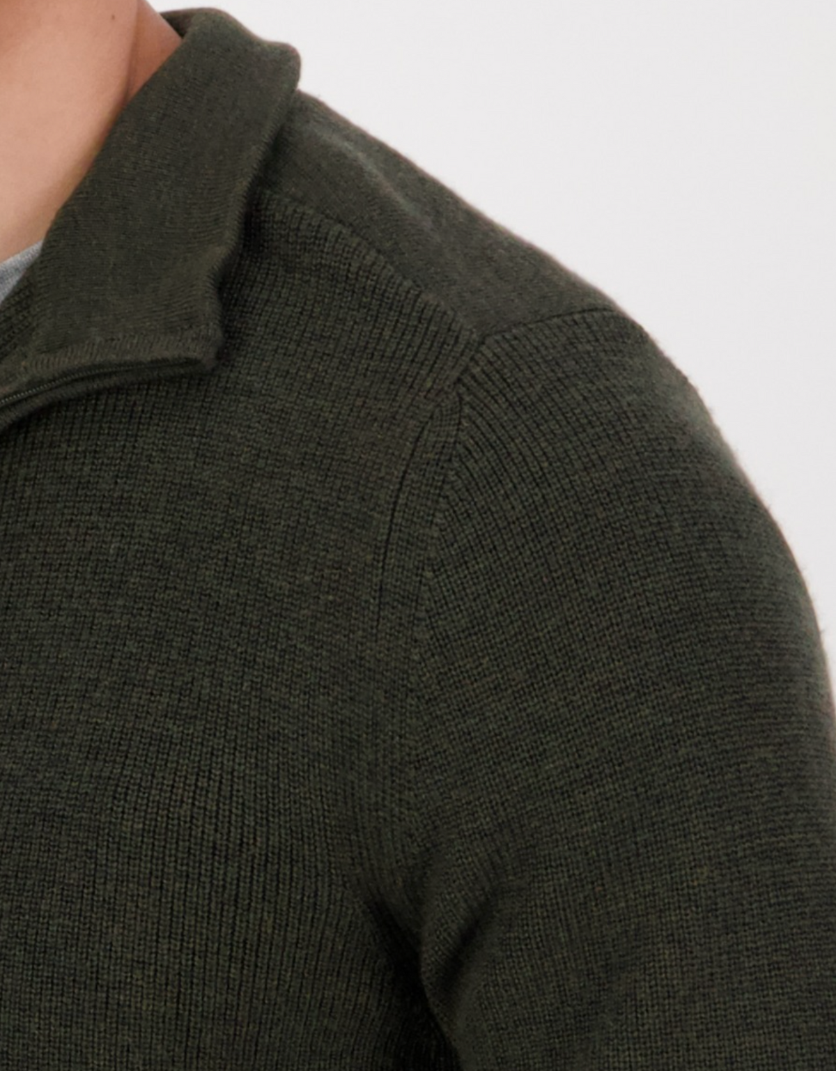 A close-up of a person wearing the Swanndri Magnet Bay v2 Merino Jumper, featuring a dark green knit with a half zip and high collar. The focus is on the Merino wool texture and stitching against a plain, light-colored background.