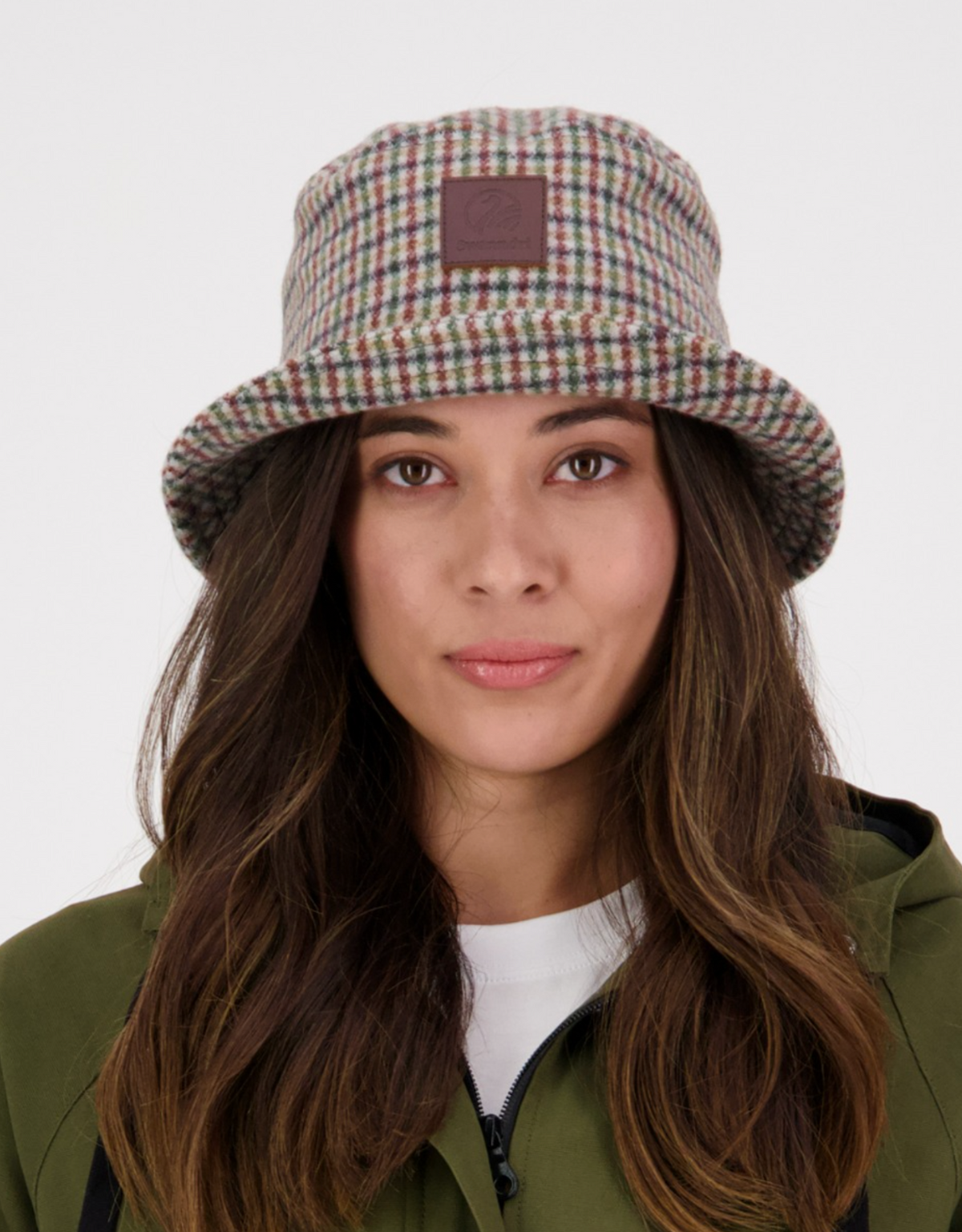 A woman wearing the Swanndri Kaimai Wool Bucket Hat with a brown patch, an olive green jacket, and a white shirt, looks directly at the camera against a plain background.