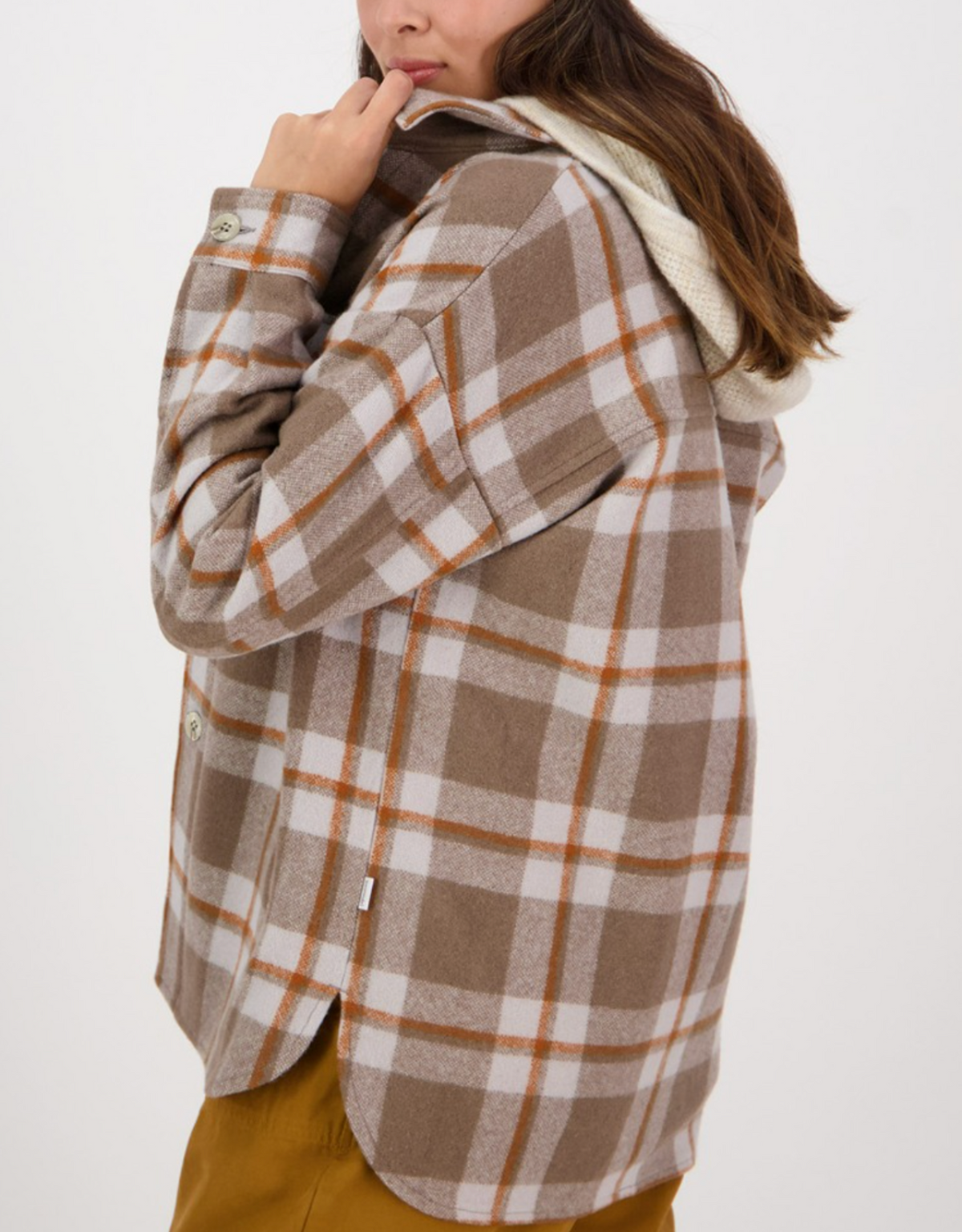 A person with long hair is shown from the back, wearing a Swanndri Anchorage Wool Shirt Jacket in beige, white, and orange plaid, layered over a white hoodie. The person appears to be holding the front of the jacket's collar with their left hand. They are also wearing mustard-colored pants.