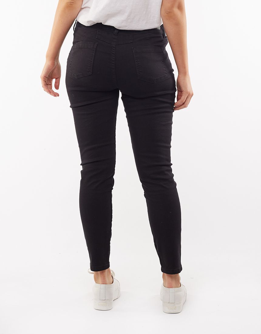 Rear view of a person standing against a white background, showcasing the Foxwood City Jean – high-waisted black skinny jeans in a cotton blend. They are wearing a white shirt tucked into the jeans and white sneakers. The jeans feature back pockets and a fitted silhouette.