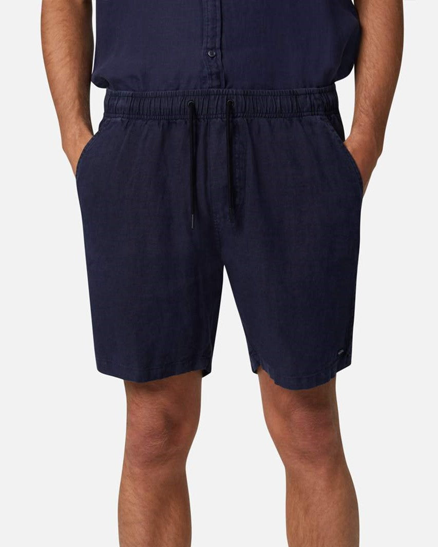 A person models Industrie The Marina Linen Short in navy, hands in pockets, highlighting its elastic drawstring waist for comfort. The plain white background accentuates the casual appeal of these comfortable shorts.