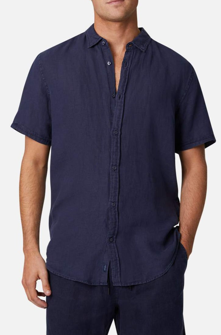A man wearing the Industrie The Tennyson Linen Short Sleeve Shirt in dark blue, featuring a relaxed fit with short sleeves and an open collar, stands against a plain background. With his left hand in his pocket, he exudes effortless style.