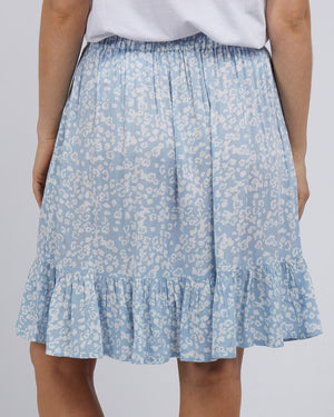 A person showcases the back view of a stylish ensemble pairing a white top with the Foxwood Kina Skirt, featuring an elastic waist for comfort, in blue with a light floral pattern and a ruffled hem.