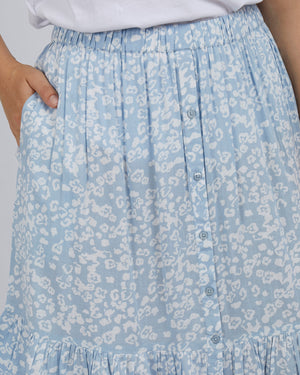 A person wears the Foxwood Kina Skirt, a 100% viscose blue skirt with a white floral pattern, elastic waist, front buttons, and ruffled bottom. They pair it with a white top and have one hand in the skirt's pocket.
