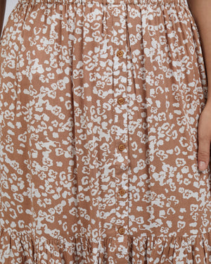 A close-up shows the Foxwood Kina Skirt, a brown piece with a white abstract floral pattern. It features a row of small buttons down the center, gathered details at the elastic waist, and a ruffle hem. A hand gently touches this elegant 100% Viscose garment.