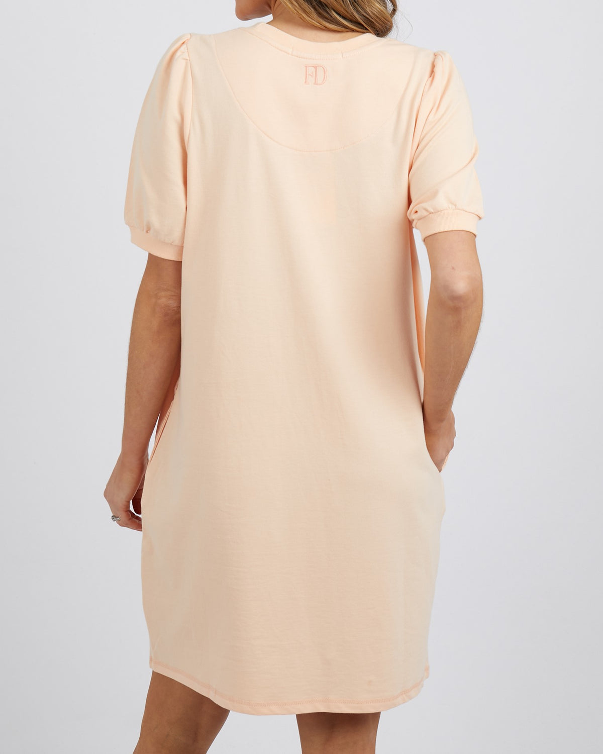 A woman is seen from the back wearing the Foxwood Zaliah Dress, a loose-fit, short-sleeved peach dress made from 100% cotton. Her left hand rests in her pocket and her long hair falls partially visible against a plain background.