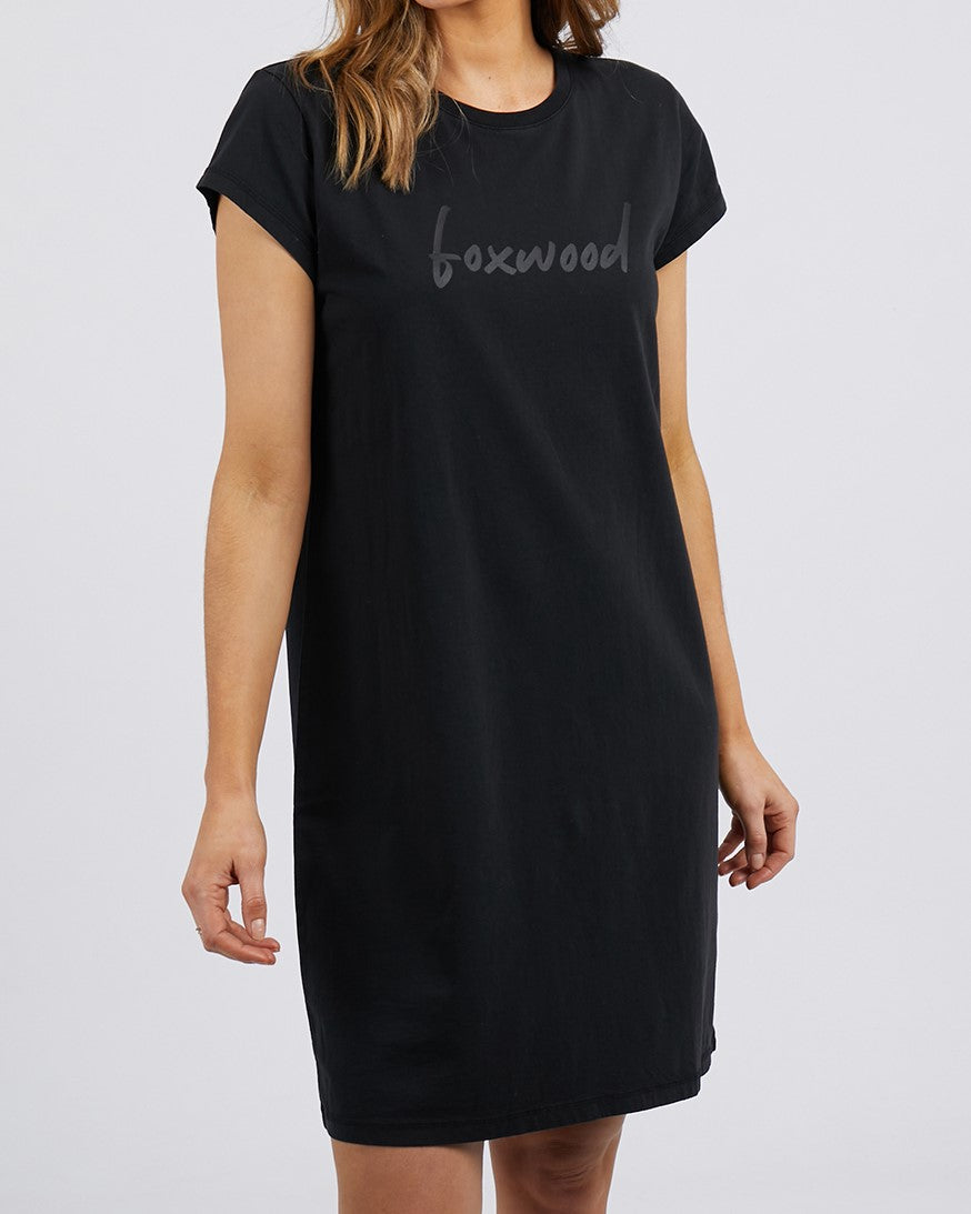 Someone is wearing the Foxwood Signature Dress, a comfy 100% cotton black t-shirt dress with short sleeves and an oversized fit. The word "boxwood" in gray is printed on the chest against a plain gray background, ideal for relaxed days.