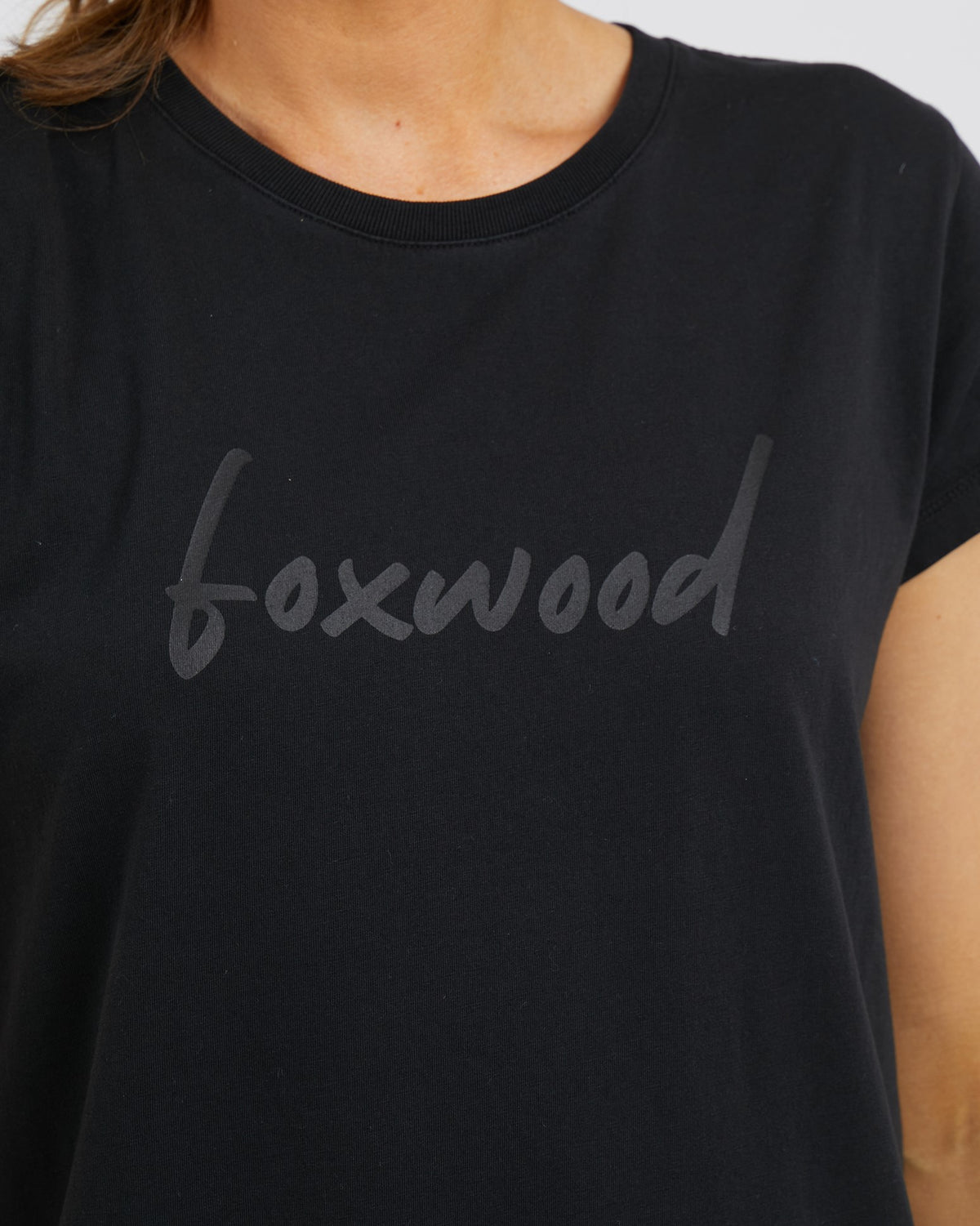 A person wears the Foxwood Signature Dress, featuring "foxwood" in lowercase gray letters on a black t-shirt. The image highlights its sleek design, oversized fit for comfort, and 100% cotton fabric.