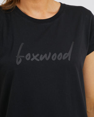 A person wears the Foxwood Signature Dress, featuring "foxwood" in lowercase gray letters on a black t-shirt. The image highlights its sleek design, oversized fit for comfort, and 100% cotton fabric.