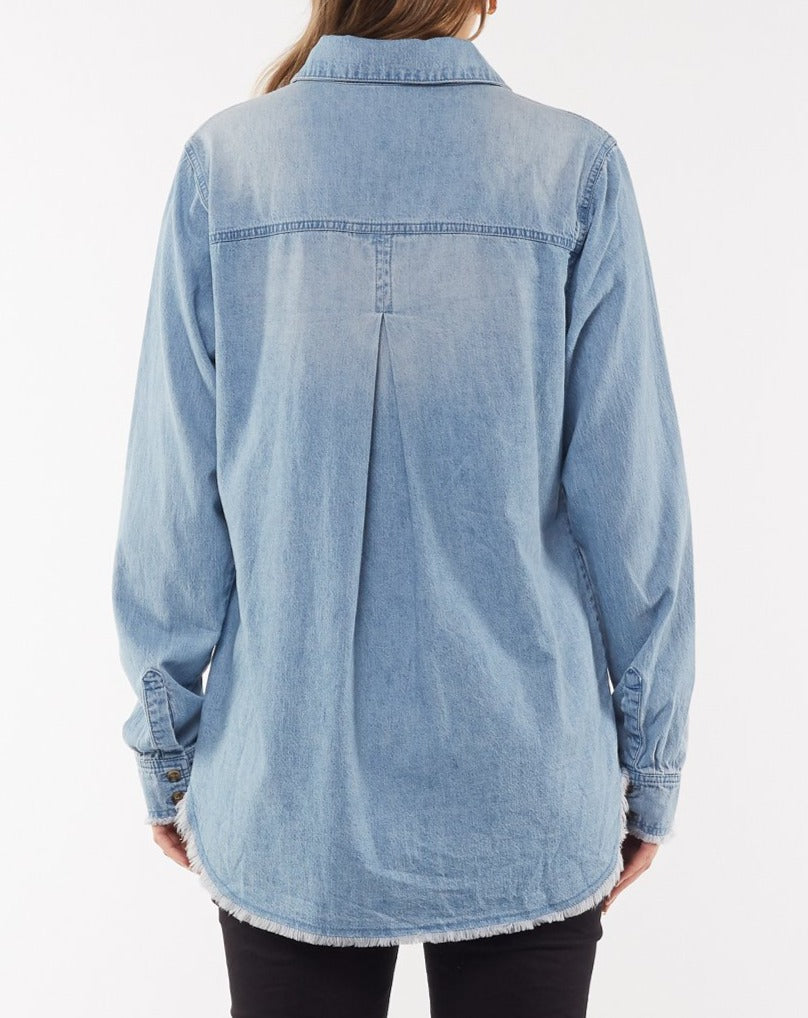 A person wearing the Foxwood Noah Shirt in light blue denim is seen from the back. The cotton shirt features buttoned cuffs, a yoke with a central pleat, and a button front. The hem of the shirt is frayed, and the person is also wearing black pants.