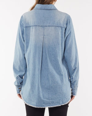 A person wearing the Foxwood Noah Shirt in light blue denim is seen from the back. The cotton shirt features buttoned cuffs, a yoke with a central pleat, and a button front. The hem of the shirt is frayed, and the person is also wearing black pants.