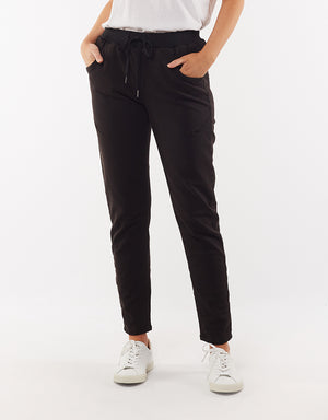 A person wearing black Foxwood Sylvia Jogger pants, a white T-shirt, and white sneakers stands against a plain white background. The image captures them from the neck down, showcasing the comfortable jogger pants with hands in their pockets.