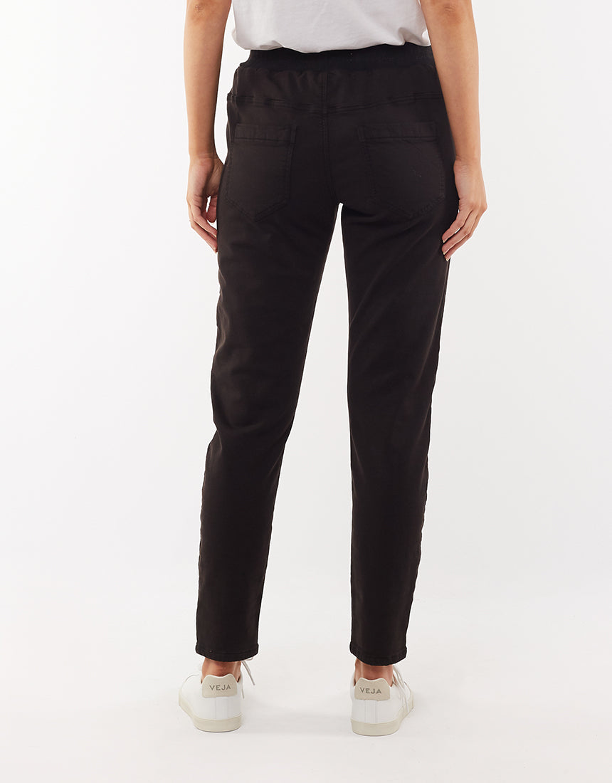 A person standing and facing away from the camera is dressed in a white cotton elastane shirt paired with high-waisted black "Foxwood Sylvia Jogger" pants. The individual, with their arms relaxed at their sides, is also sporting white sneakers against a plain, white background.
