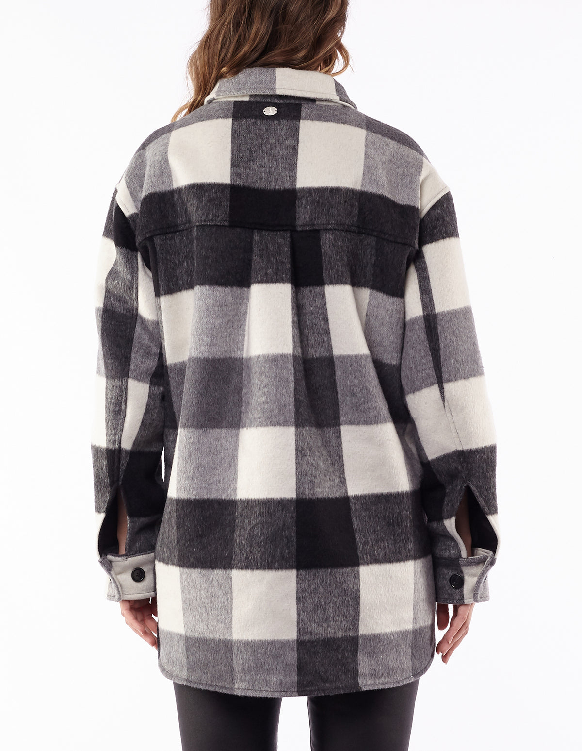 A woman with loose brown hair faces away, wearing the Foxwood Lyon Shacket—featuring black, white, and gray plaid, buttoned cuffs, and a rounded hem—styled with black pants.