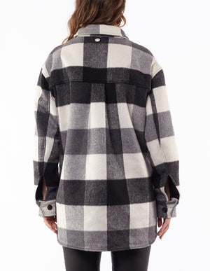 A woman with loose brown hair faces away, wearing the Foxwood Lyon Shacket—featuring black, white, and gray plaid, buttoned cuffs, and a rounded hem—styled with black pants.