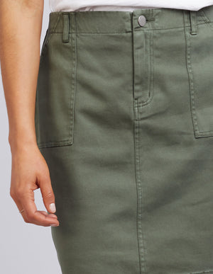 A person stands calmly in an olive green Foxwood Charlee cotton skirt with large front pockets and a white top. The skirt, secured by a button at the waistband, elegantly complements their outfit as their left hand rests effortlessly by their side.