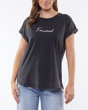 A person confidently wears rolled-up short sleeves of a "Foxwood Signature Tee" in black, crafted from lightweight, breathable fabric. They pair it with light blue jeans and stand against a plain white backdrop, one hand on their hip.