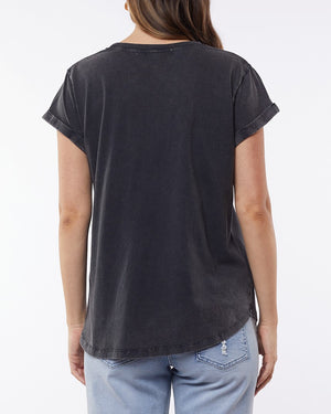 View of a person with long hair wearing the Foxwood Signature Tee, a faded black T-shirt made from lightweight breathable material, paired with light blue jeans. The background is plain.
