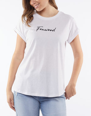 A woman models the Foxwood Signature Tee, a lightweight white shirt with "Foxwood" in cursive. She pairs it with light blue jeans, smiling against a plain white backdrop with her shoulder-length hair and one hand in her pocket.