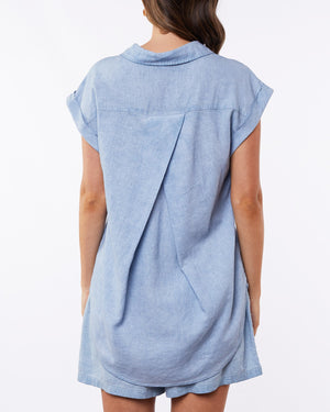 The image showcases the Foxwood Palm Shirt, a light blue short-sleeve denim dress with a relaxed fit and pleated back detail, falling just below the knees. The stylish piece pops against a plain white background.