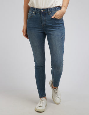 A person in a white top and Foxwood City Jean, standing slightly apart. They wear white sneakers and rest their left hand on their hip, against a plain gray background.