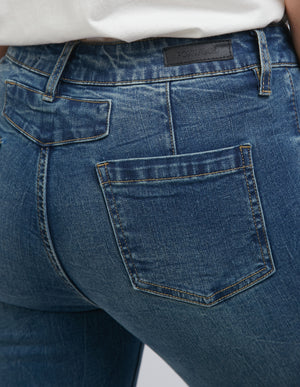 Close-up of a person wearing the high-waist Foxwood City Jean, featuring faded cotton design, stitching details, back pocket, belt loops, and skinny leg fit. A black logo tag is visible on the waistband of these blue jeans paired with a white top.