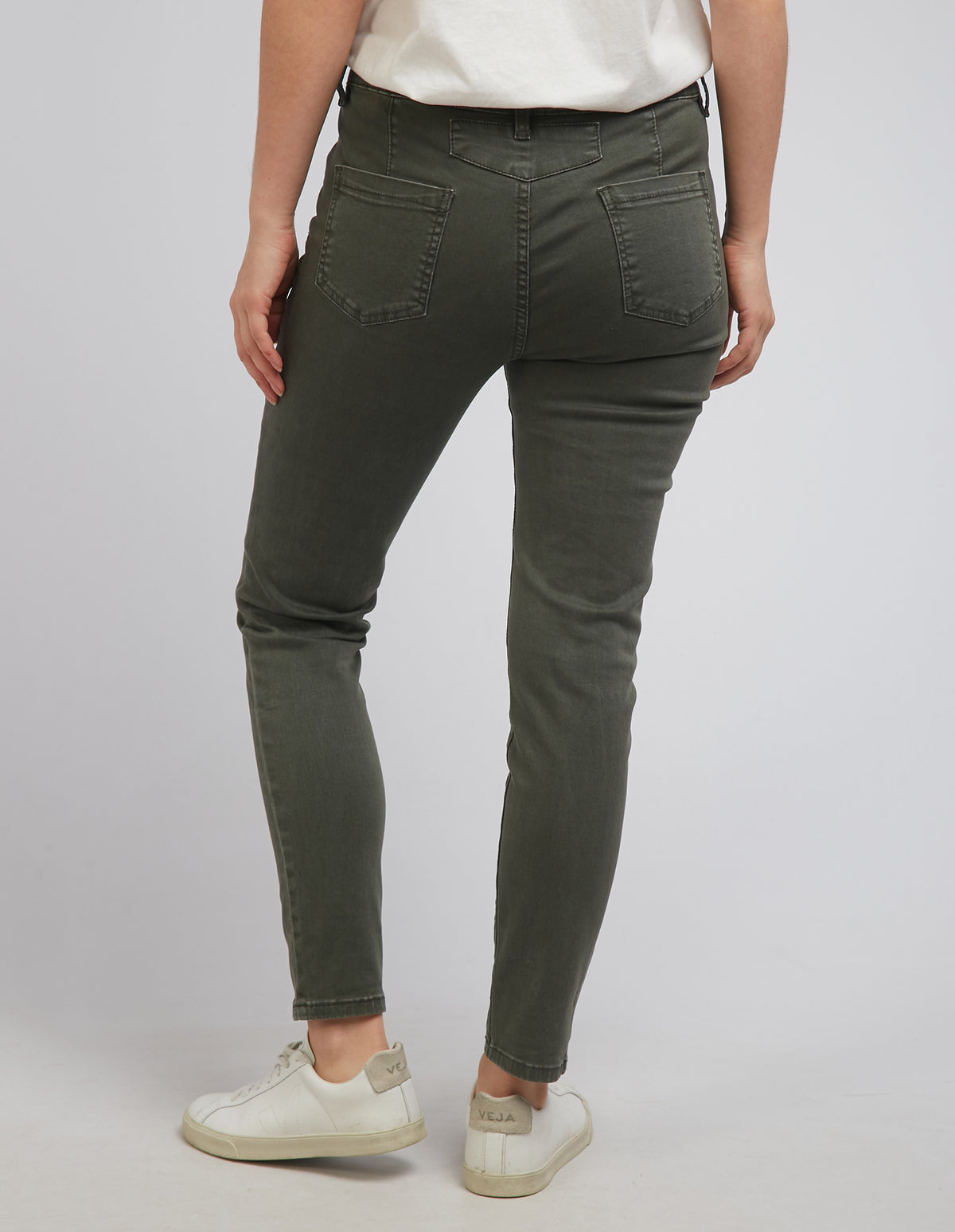 A person in high-waisted Foxwood City Jeans, olive green, and white sneakers, is photographed from behind. They stand on a light gray surface with a pale gray background.